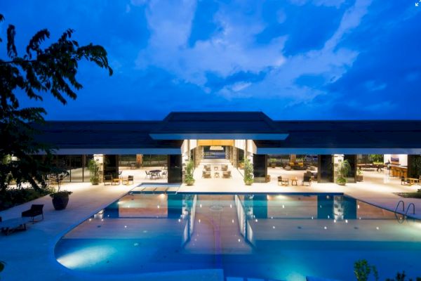 A luxurious pool area at night, with a modern building in the background, under a blue, cloudy sky, and trees partially visible.