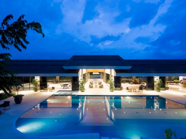 A luxurious pool area at night, with a modern building in the background, under a blue, cloudy sky, and trees partially visible.
