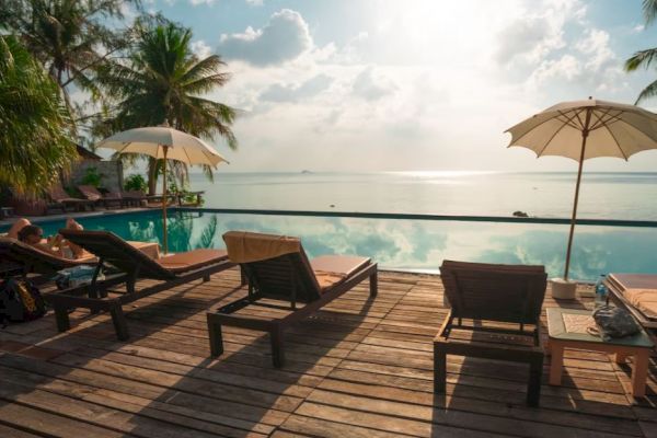 A serene poolside setting with lounge chairs, umbrellas, palm trees, and a peaceful view of the ocean under a partly cloudy sky.