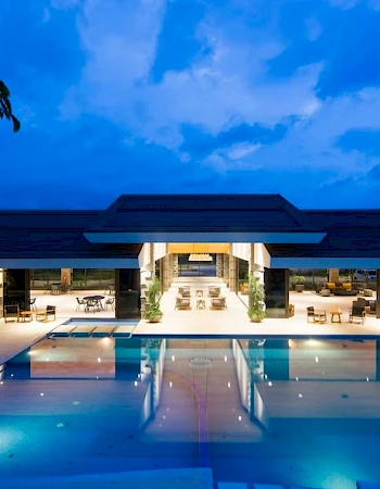 A modern building with expansive windows overlooks a large, illuminated swimming pool at dusk, surrounded by seating arrangements.