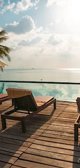 A tropical poolside scene with lounge chairs, umbrellas, and palm trees overlooks a serene body of water under a sunny sky.