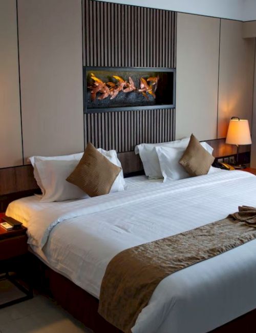 A cozy hotel room with a large bed, decorative pillows, side lamps, a modern fireplace, and curtains by a window, offering a relaxing ambiance.
