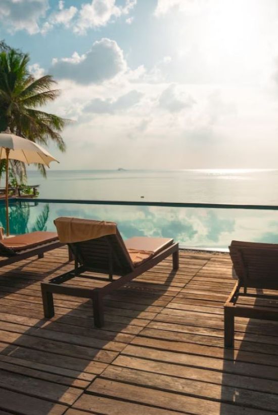 A serene poolside setting with lounge chairs, umbrellas, palm trees, and a view of the ocean under a bright sky.