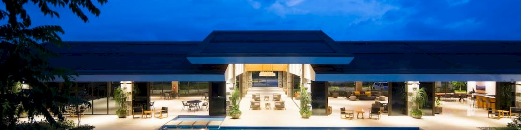 The image shows a luxurious house with a large, illuminated pool in front, set against a deep blue sky at dusk.