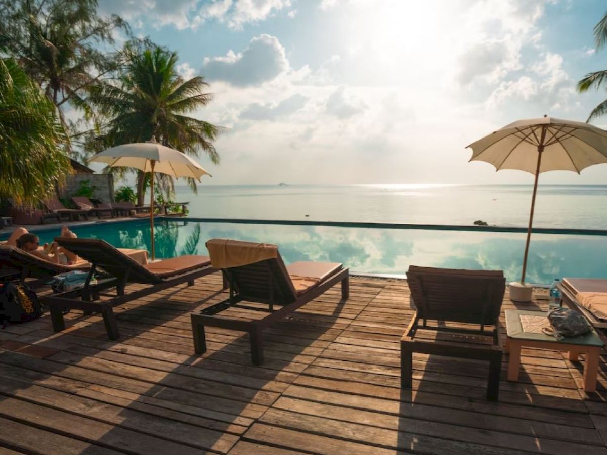 Wooden deck with loungers and umbrellas by a pool; calm water and distant horizon with clouds and palm trees in the background.