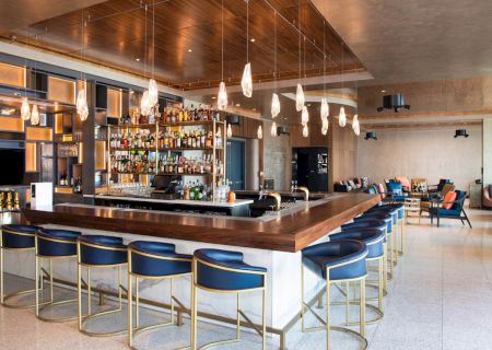 A modern bar with a sleek design features blue stools, hanging lights, and a fully stocked liquor shelf, creating a trendy atmosphere.