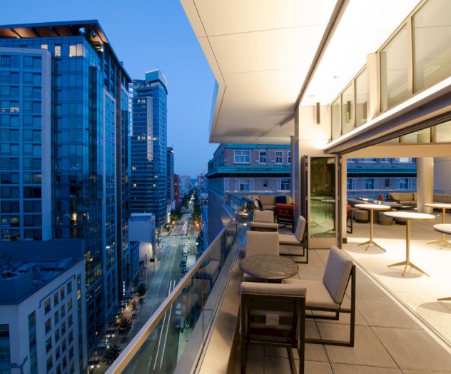Cityscape view from a modern balcony at dusk, featuring tall buildings and a seating area with tables and chairs.