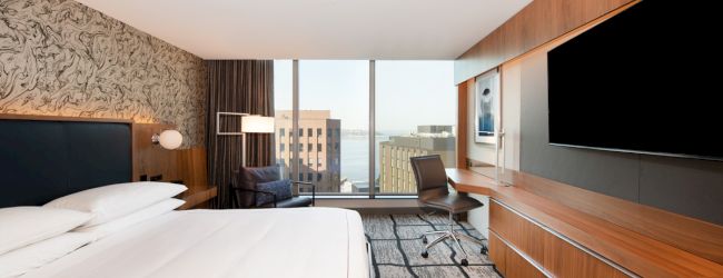 A modern hotel room features a large bed, wall-mounted TV, desk, and chair, with a window view of buildings and a body of water.
