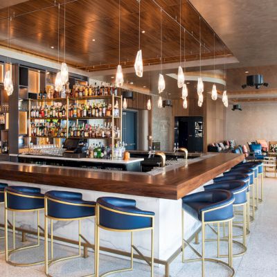 A modern bar with blue seating, warm lighting, and a well-stocked shelf. Lounge area with sofas is visible in the background.