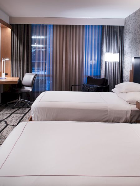 The image shows a modern hotel room with two beds, a desk, a large TV, decorative lighting, and patterned carpeting and wall design.