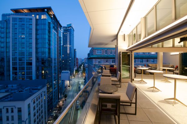 A modern cityscape at dusk is visible from a balcony with tables and chairs, overlooking tall buildings and a street below.