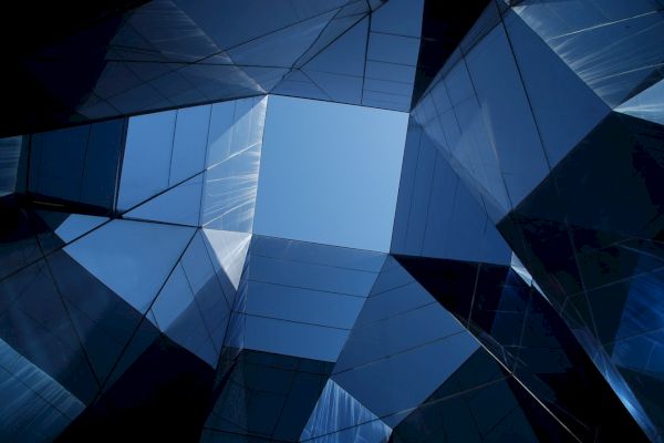 The image shows an upward view of modern glass buildings with reflective surfaces, creating a geometric pattern against the sky.