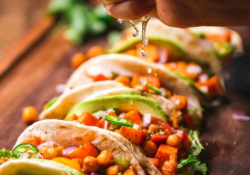 A hand is squeezing lime over a row of soft tacos filled with chickpeas, diced vegetables, cilantro, and avocado slices on a wooden surface.