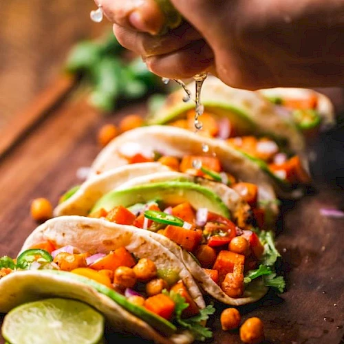 A hand is squeezing lime over a row of soft tacos filled with chickpeas, diced vegetables, cilantro, and avocado slices on a wooden surface.