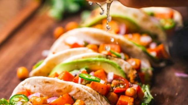 A hand is squeezing lime over a row of soft tacos filled with chickpeas, diced vegetables, cilantro, and avocado slices on a wooden surface.