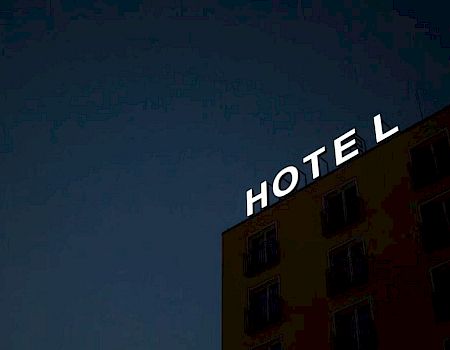 A building facade at night with a glowing "HOTEL" sign missing the letter "L," against a dark sky.