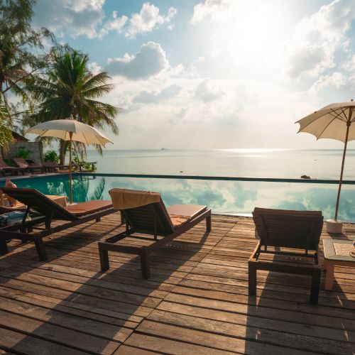 Lounge chairs and umbrellas by an infinity pool overlooking the ocean, with palm trees and a bright sky in the background.
