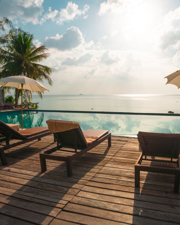 Lounge chairs and umbrellas by an infinity pool overlooking the ocean, with palm trees and a bright sky in the background.