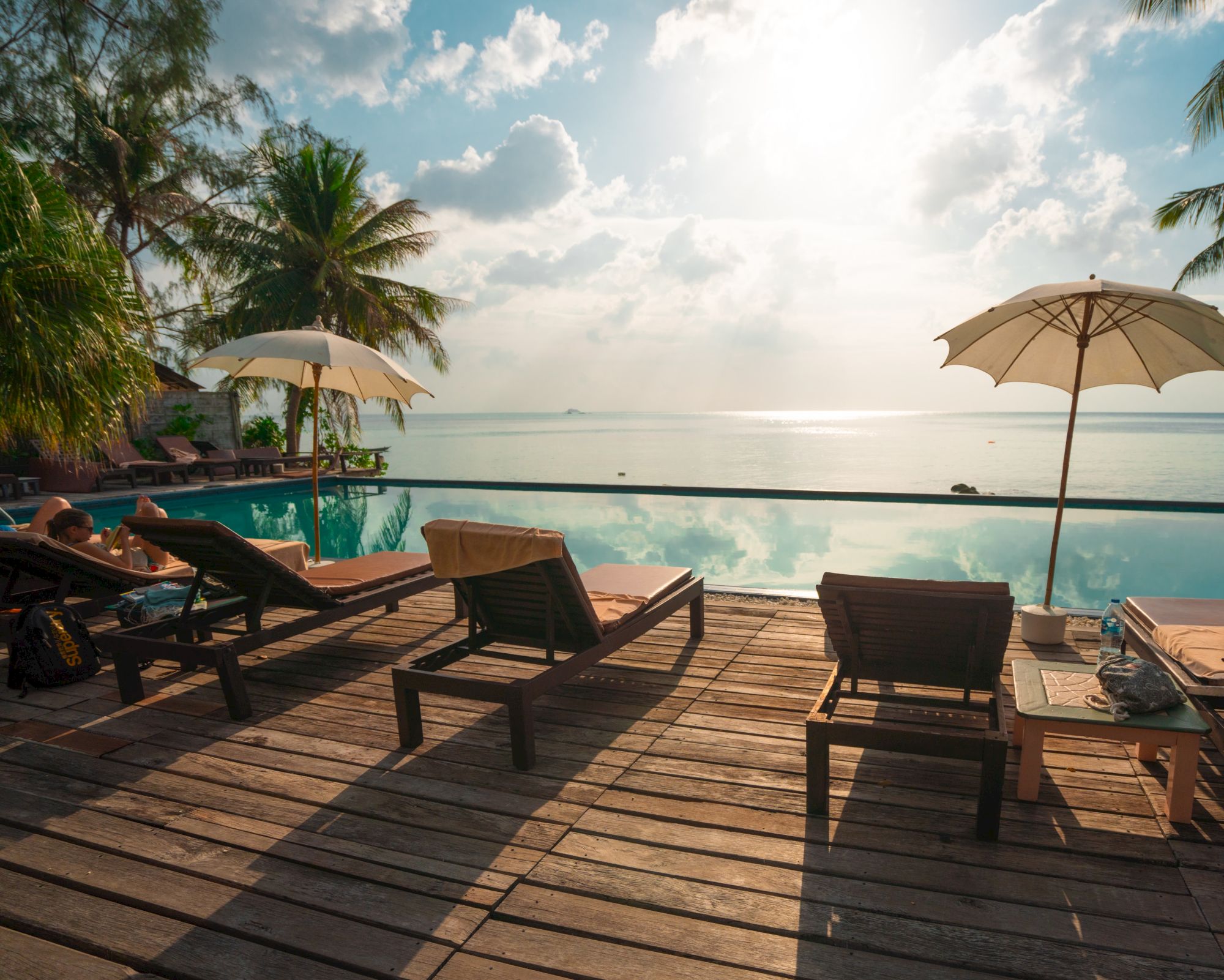 A serene tropical setting with lounge chairs, umbrellas, and a pool overlooking the ocean, framed by palm trees and a bright sky.