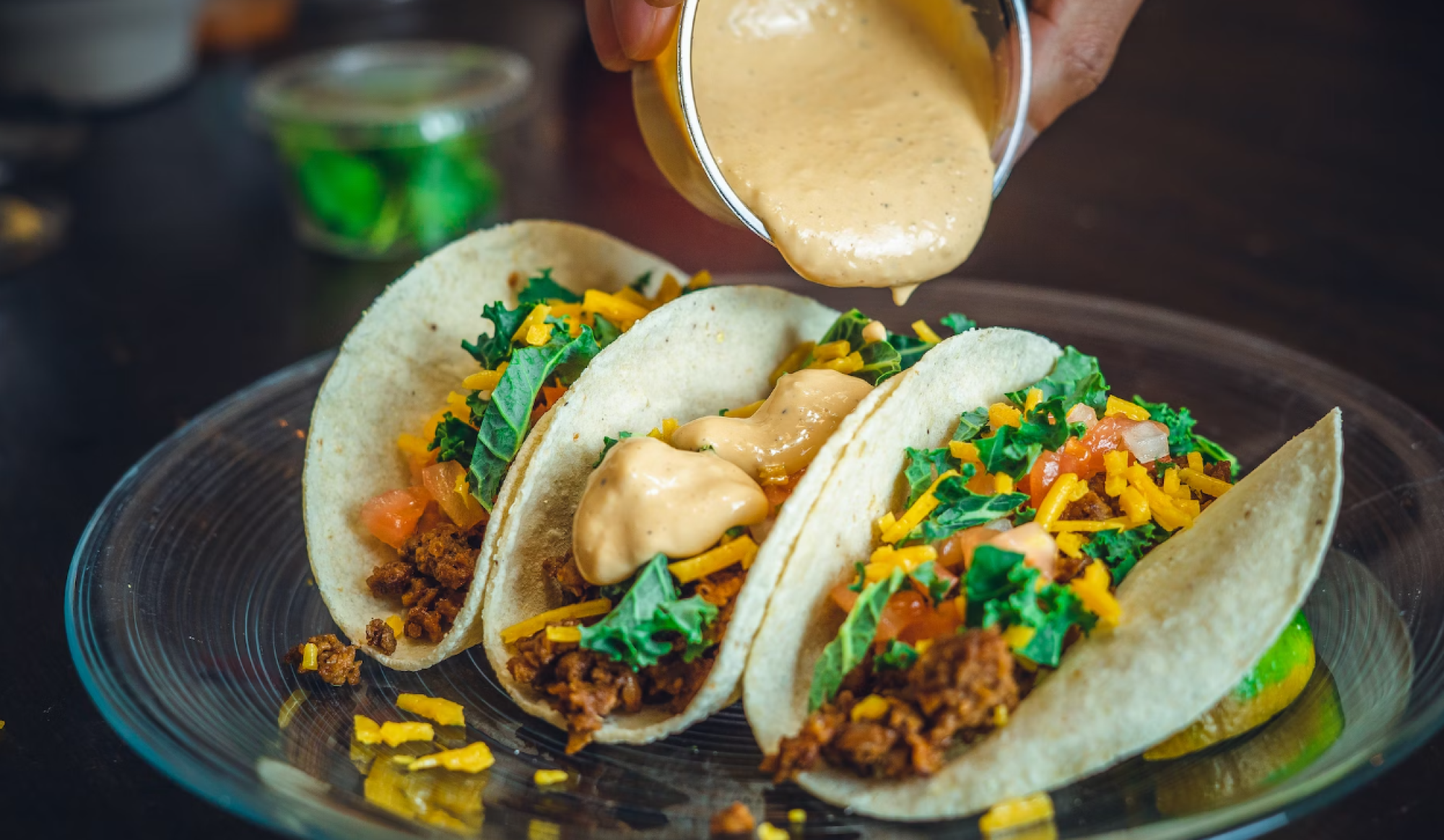 Three tacos with ground filling, cheese, lettuce, and sauce being poured on top, on a clear plate.