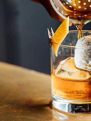A drink being poured into a glass with ice and a citrus peel garnish, placed on a bar counter.