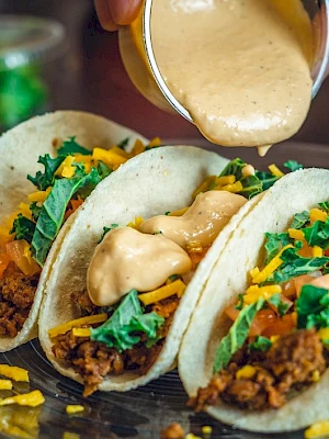 Three tacos with ground meat, shredded cheese, lettuce, tomato, and sauce being poured on top.
