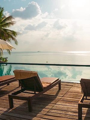 A tranquil poolside setting with lounge chairs, umbrellas, trees, and a sea view under a cloudy sky.