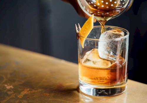 A glass with ice and a citrus peel garnish, being filled with a brown liquid using a cocktail strainer.
