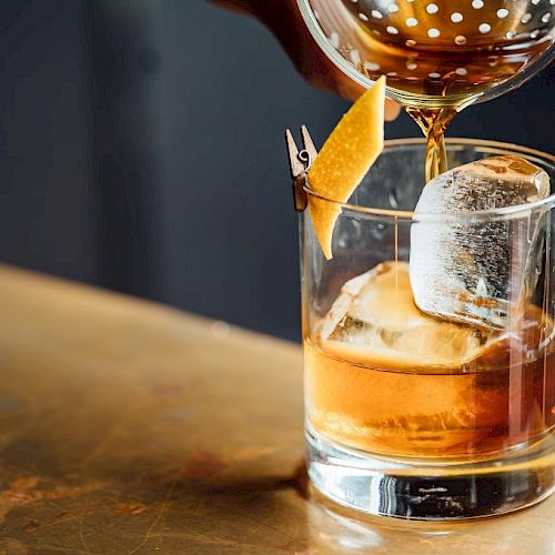 A glass with ice and a citrus peel garnish, being filled with a brown liquid using a cocktail strainer.