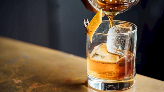 A glass with ice and a citrus peel garnish, being filled with a brown liquid using a cocktail strainer.