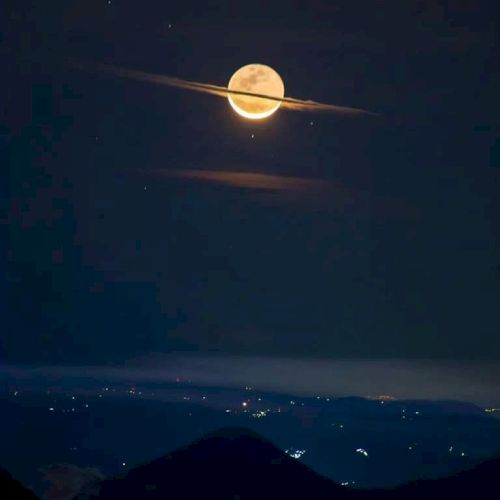 A full moon appears with clouds creating an illusion of Saturn's rings over a landscape with distant city lights, against a dark sky.