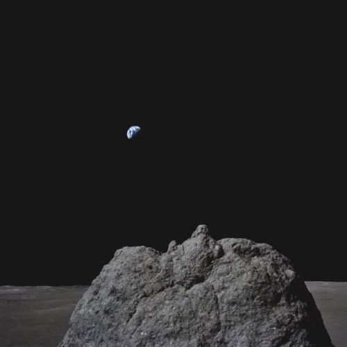 A lunar surface with a prominent rock in the foreground and Earth visible in the distant black sky.