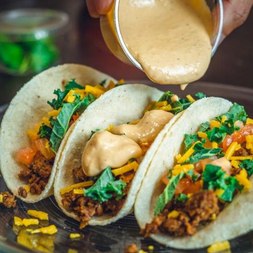 Three tacos with meat, greens, and cheese are being topped with a creamy sauce on a plate.
