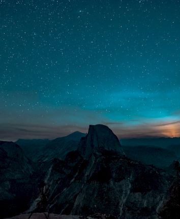 A starry night sky over a mountain range with a glowing horizon, possibly the moon rising, and dark silhouettes of trees in the foreground.