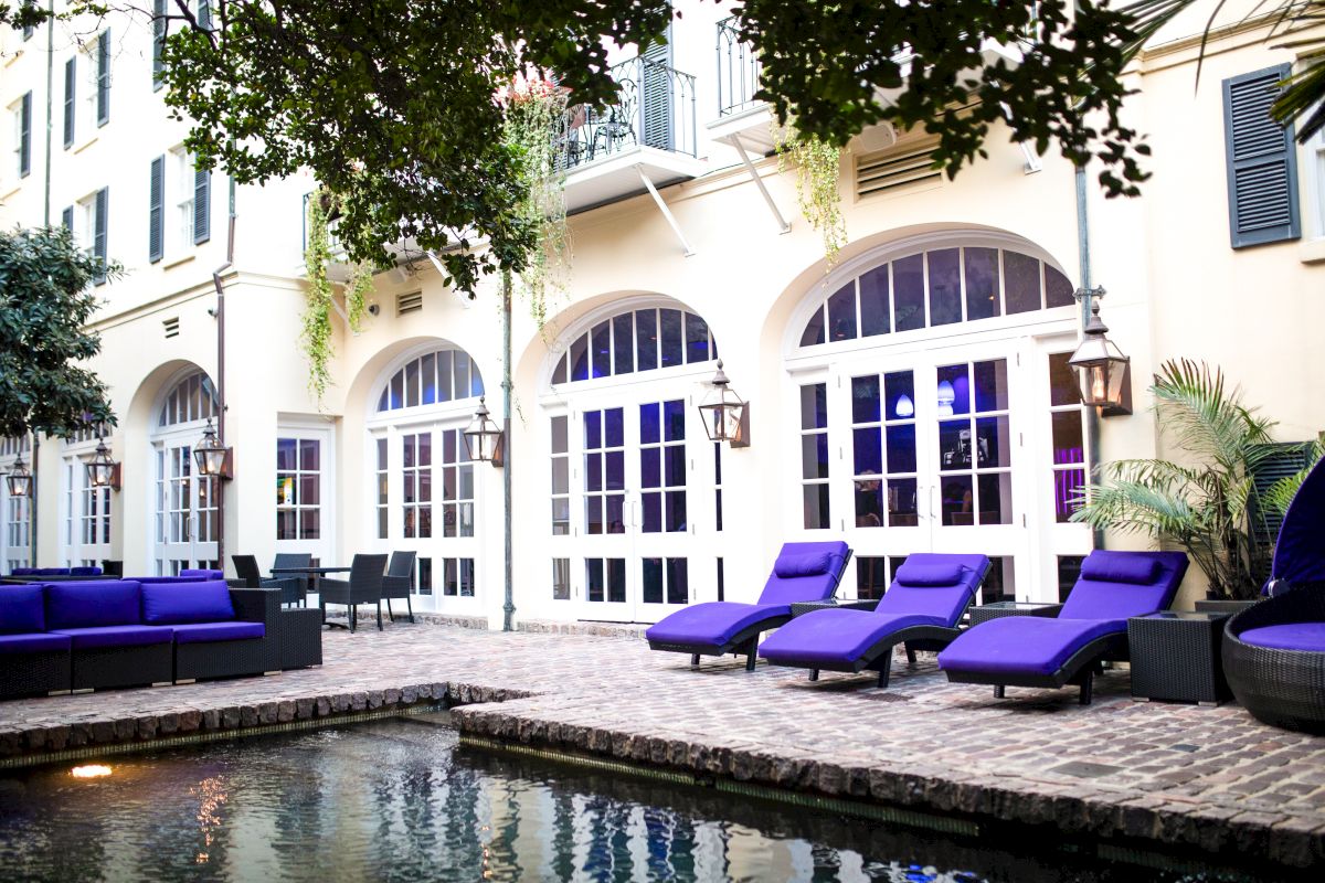 A courtyard with a pool, purple lounge chairs, a sofa, and a potted plant in front of a building with large windows.