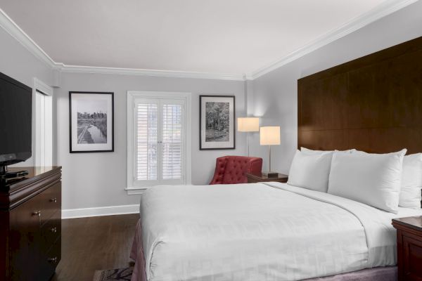 A cozy hotel room with a large bed, red chair, wooden furniture, photo frames, and a flat-screen TV. White walls and soft lighting complete the look.