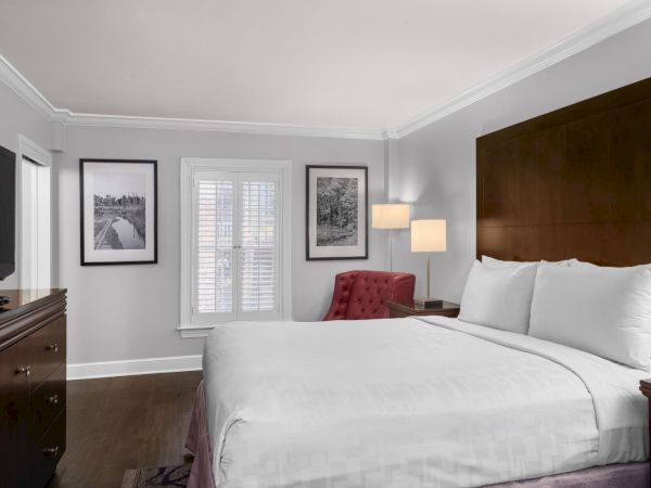 A cozy hotel room with a large bed, red chair, wooden furniture, photo frames, and a flat-screen TV. White walls and soft lighting complete the look.