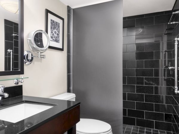 A modern bathroom with a black-tiled shower, a mirror, sink, toilet, and wall art in a sleek design.