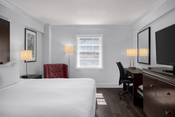 The image shows a hotel room with a bed, red chair, desk, TV, and window, all in a minimalist, modern style.