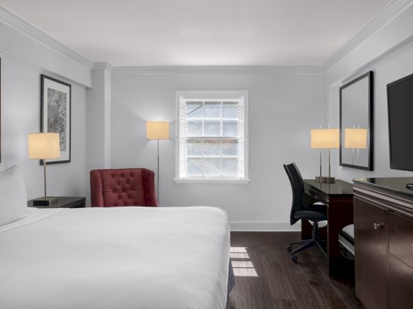 The image shows a hotel room with a bed, red chair, desk, TV, and window, all in a minimalist, modern style.