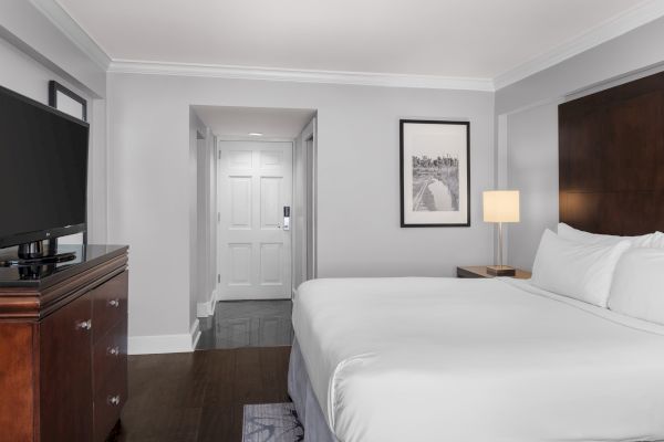 A hotel room with a bed, TV on a wooden cabinet, lamp, framed artwork on the wall, and a closed door in the background.