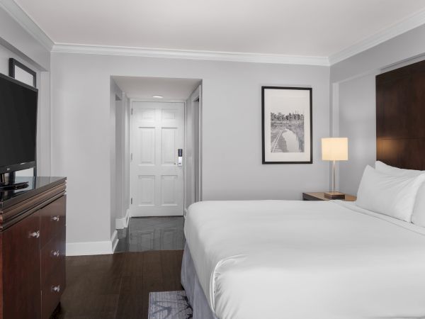 A hotel room with a bed, TV on a wooden cabinet, lamp, framed artwork on the wall, and a closed door in the background.