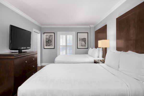 The image shows a hotel room with two beds, a TV on a dresser, framed artwork, a lamp, and a window with shutters on a gray wall.