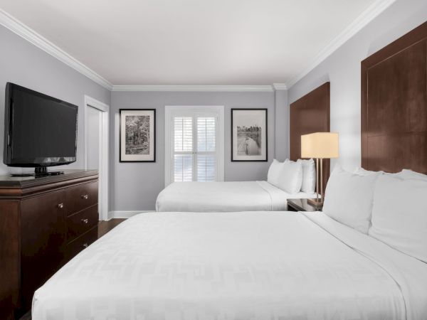 The image shows a hotel room with two beds, a TV on a dresser, framed artwork, a lamp, and a window with shutters on a gray wall.