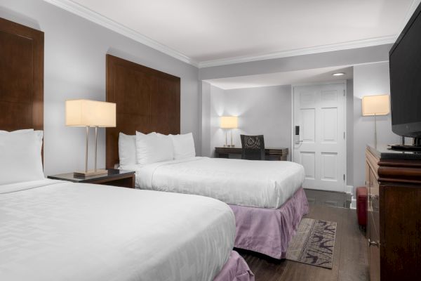 A hotel room with two beds, wooden furniture, a desk with a chair, lamps, and a TV. The decor is simple and modern.