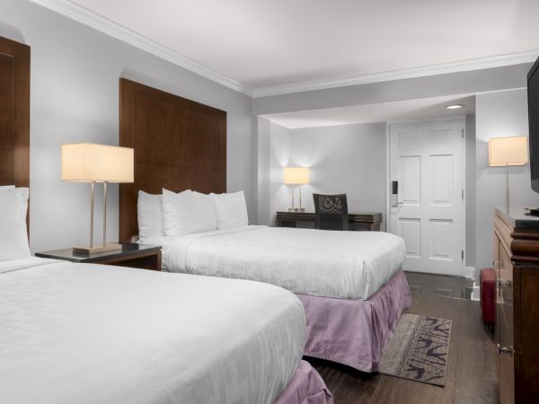 A hotel room with two beds, wooden furniture, a desk with a chair, lamps, and a TV. The decor is simple and modern.