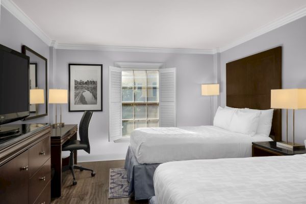 The image shows a hotel room with two beds, a desk, chair, TV, and window with shutters, all in a neutral color scheme.