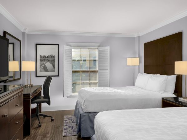 The image shows a hotel room with two beds, a desk, chair, TV, and window with shutters, all in a neutral color scheme.