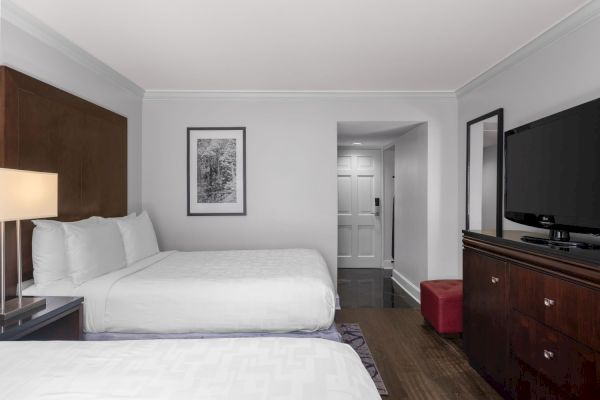 The image shows a hotel room with two beds, a TV, a lamp, framed artwork, a doorway, and a mirror.