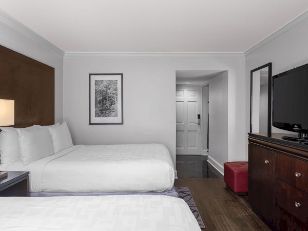 The image shows a hotel room with two beds, a TV, a lamp, framed artwork, a doorway, and a mirror.